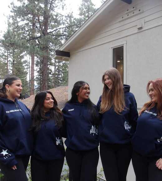 Six smiling young women in matching navy hoodies standing outdoors by a house